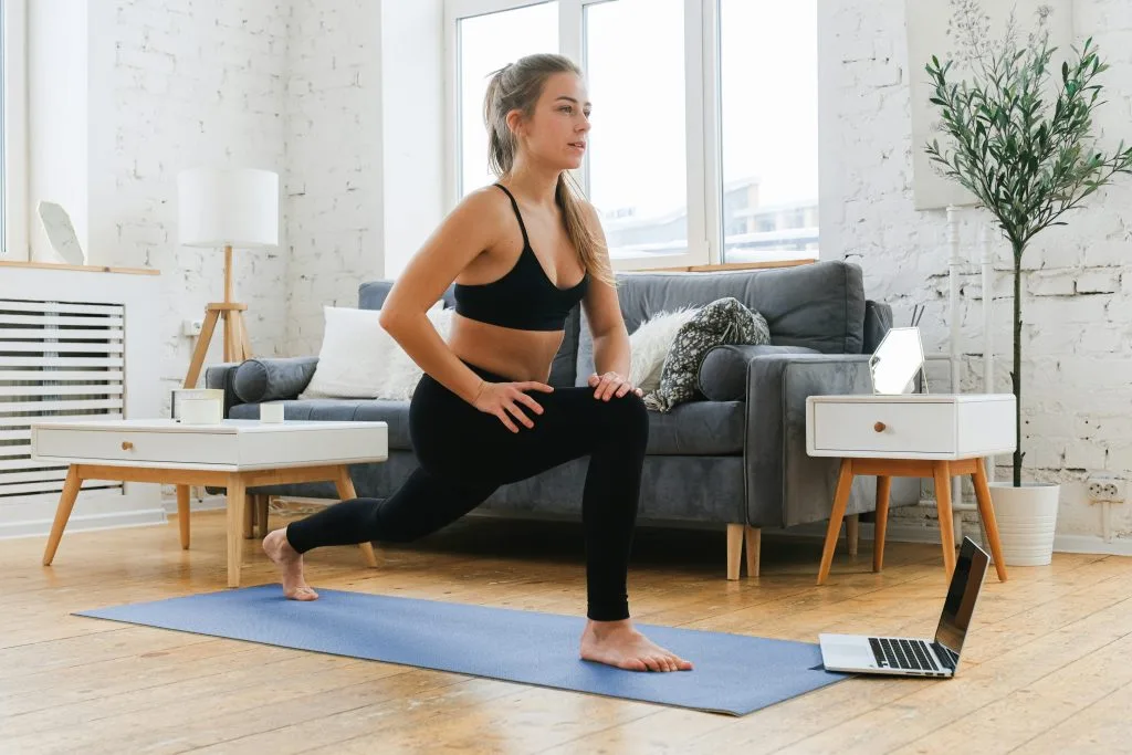 Jeune femme dans son salon en posture de la fente sur son tapis, suivant un cours en ligne de Strala Yoga gratuit sur son ordinateur portable